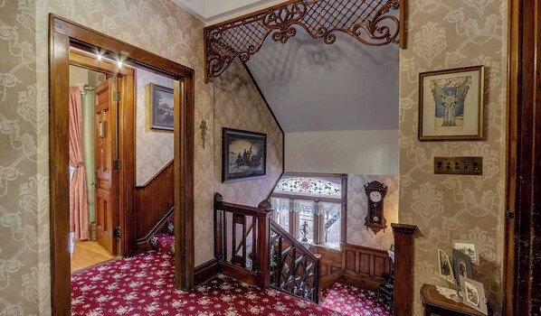 Eine Treppe mit aufwendiger Geländergestaltung und gemustertem Wandpapier, die zu einer Zwischenebene mit einer Uhr und gerahmten Bildern führt.