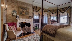 A bedroom with a four-poster bed, a fireplace, a sitting area with a chair and a sofa, and a dining area with a table and chairs. - Pias 2 Room Suite Queen Canopy bed 2nd floor (Milwaukee)