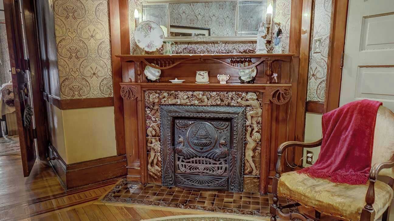 A room with a wooden fireplace, a mirror above it, and a chair with a red blanket.