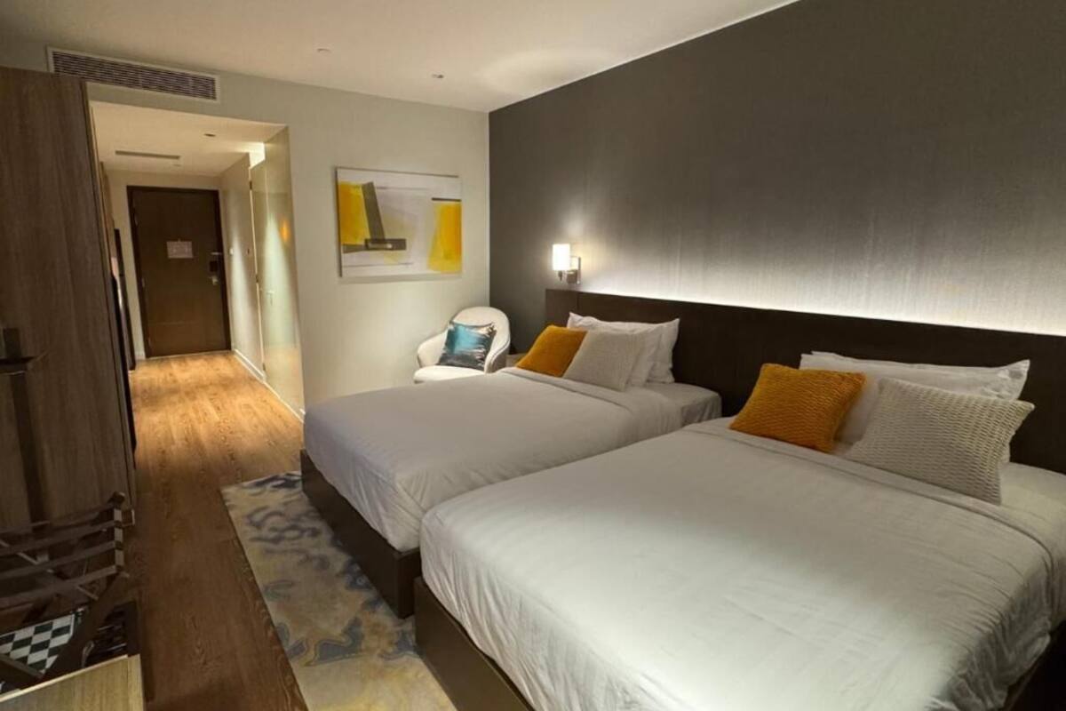 A hotel room with two beds, a wooden wardrobe, a small seating area, and a hallway leading to another room.