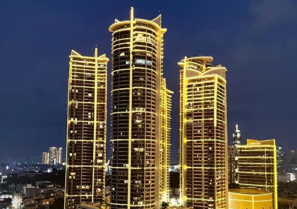 Un ensemble d’immeubles de grande hauteur, dotés de balcons illuminés, et une silhouette urbaine dense qui se dessine la nuit.
