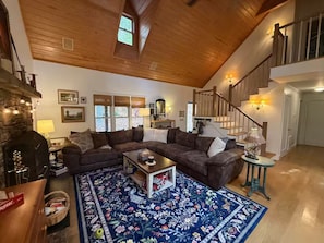 Un salon chaleureux avec un canapé d’angle, une table basse et un tapis à motifs. On y trouve une cheminée, un escalier et une fenêtre avec des stores.