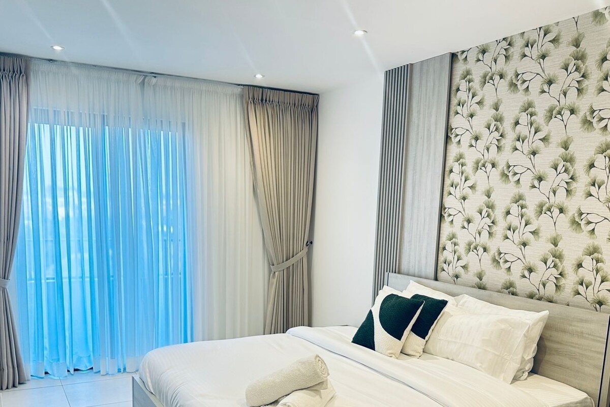 Une chambre d’hôtel moderne avec un grand lit, une tête de lit en bois et une vue sur l’extérieur à travers des rideaux transparents.