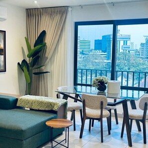 Un salon moderne avec un canapé vert, une table en verre et des chaises. Une grande fenêtre offre une vue sur la ville.
