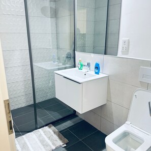 A modern bathroom with a glass shower enclosure, a white sink with a mirror above it, and a toilet. - The Bantree by Tripview Apartments (Accra)