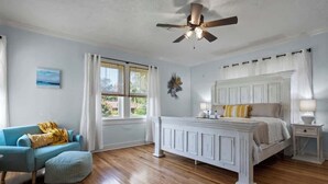 Un dormitorio con una cama blanca, un sillón azul, un ventilador de techo y una ventana con cortinas.