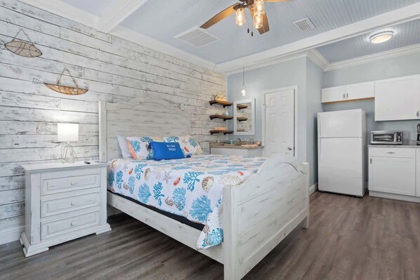 Une chambre à coucher comprenant un lit, une table de chevet, une commode, un ventilateur de plafond, et un coin cuisine avec un réfrigérateur, un four à micro-ondes et des placards.
