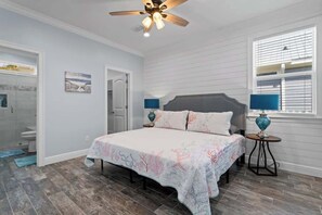 Quarto com uma cama, duas mesas de cabeceira, um ventilador de teto, janela com persianas e porta para um banheiro.
