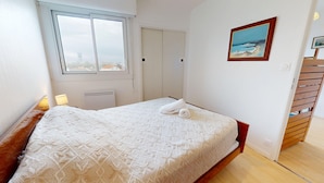 Un lit bien fait, recouvert d’une couvre-lit à motifs et agrémenté d’une serviette roulée, une fenêtre donnant sur des immeubles, une porte et un tableau accroché au mur.