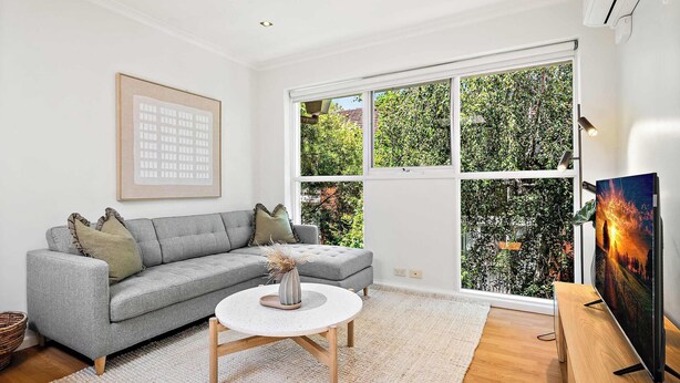 A modern living room with a grey sectional sofa, a round coffee table, and a large window with a view of greenery. - South Yarra Apt | A/C, Parking & Easy Transport (South Yarra)