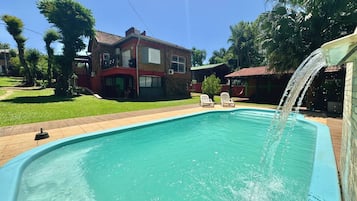 Una piscina con cascada, rodeada de un césped cuidado y una casa de dos pisos con techo rojo.