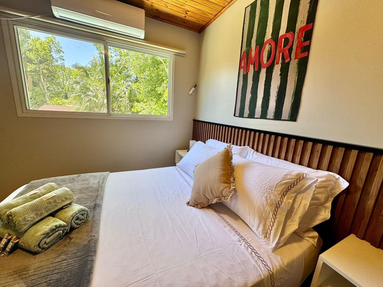 Una cama bien hecha con sábanas blancas y un cojín decorativo, una ventana con vistas a la vegetación y una pared con una obra de arte a rayas.