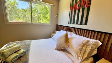 Una cama bien hecha con sábanas blancas y un cojín decorativo, una ventana con vistas a la vegetación y una pared con una obra de arte a rayas.