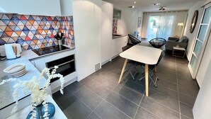 A modern kitchen with a tiled backsplash, a dining area with a white table and chairs, and a living area with a sofa and a small side table.