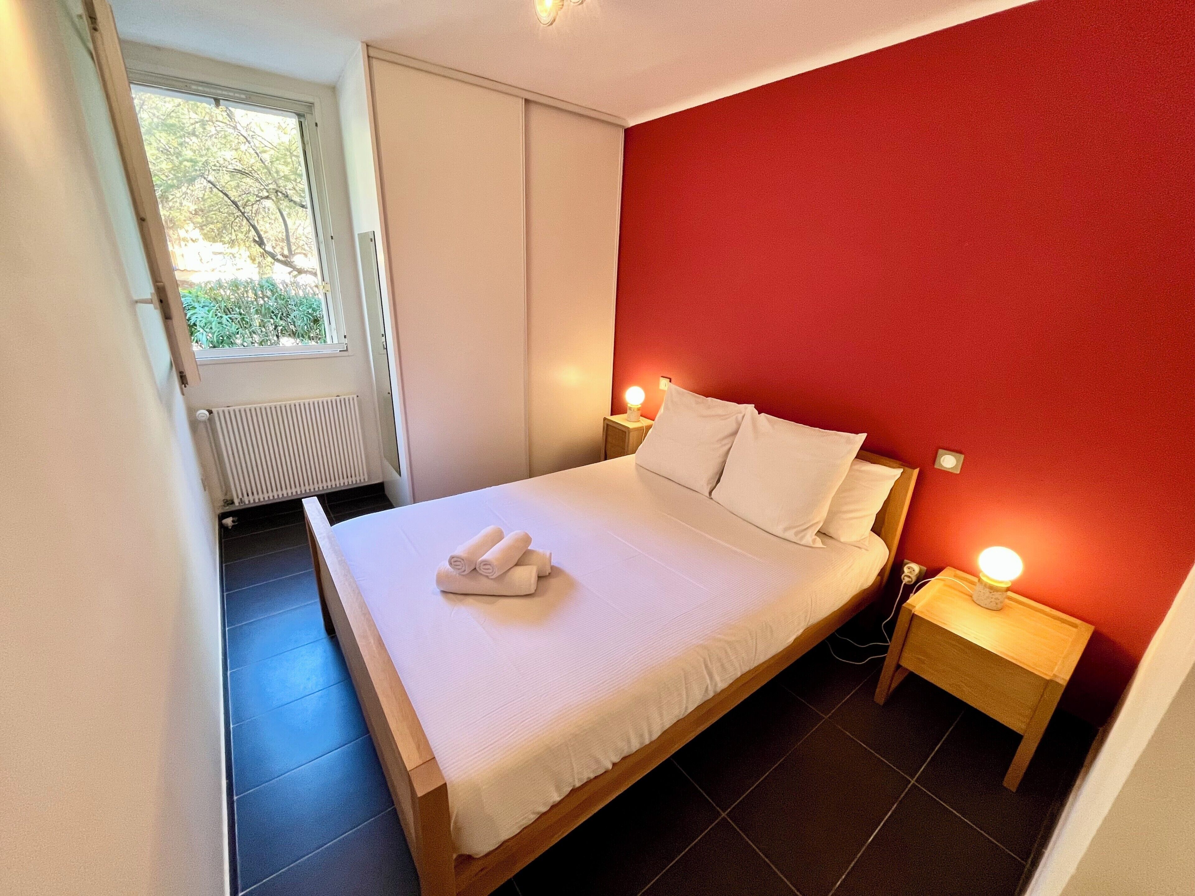Een net opgemaakt bed met witte lakens en kussens, een nachtkastje met een lamp, en een raam met gordijnen.