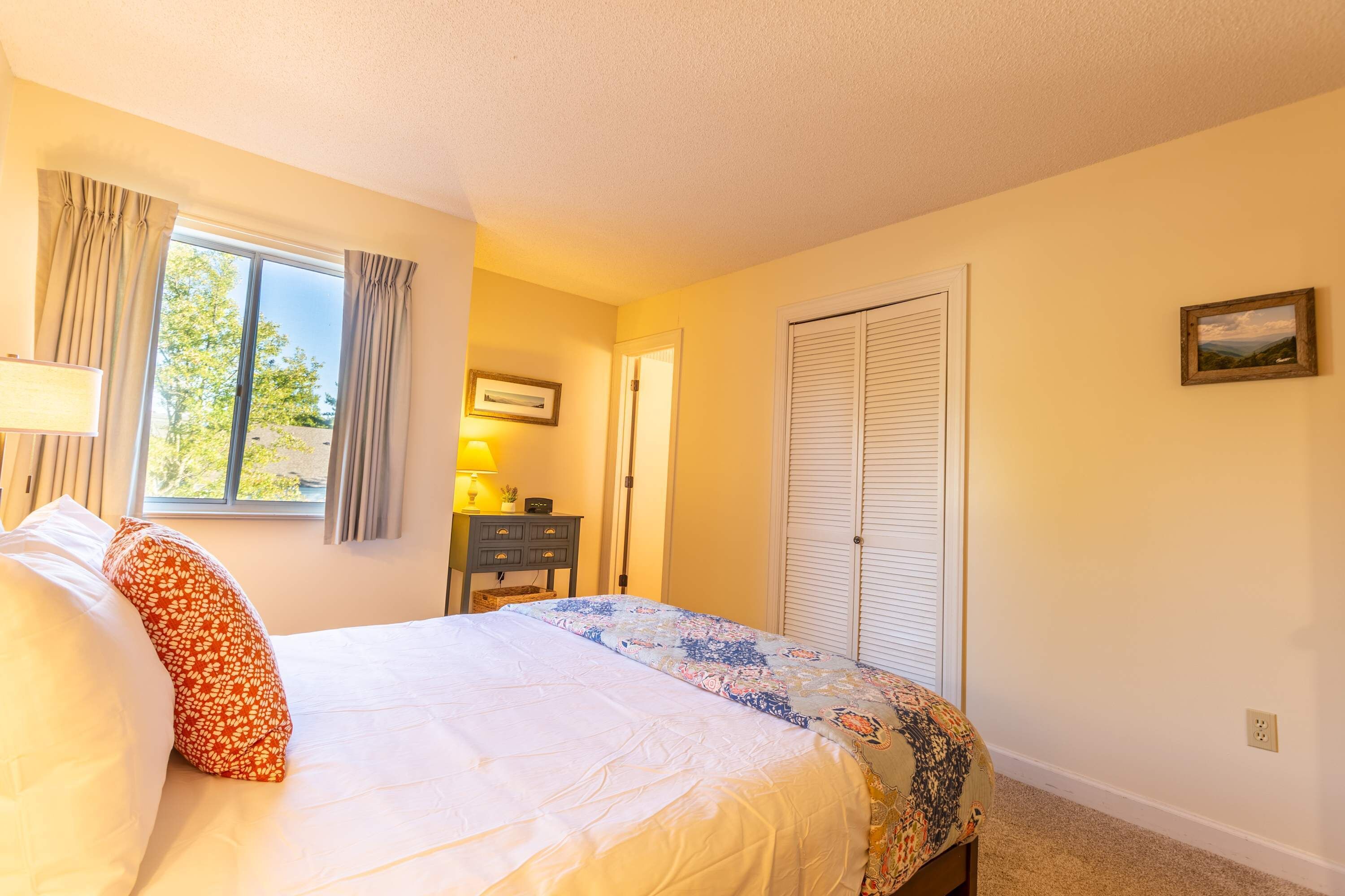 A bedroom with a bed, a nightstand, a lamp, a window with curtains, a door, and a framed picture on the wall.