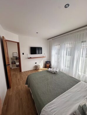 Ein Schlafzimmer mit Bett, Wandfernseher, Holzboden, Fenster mit durchsichtigen Vorhängen und einem kleinen Nachttisch mit Handtüchern.