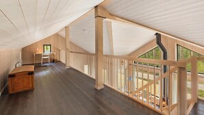 Une pièce mansardée spacieuse avec un plafond haut, des poutres en bois, un bureau et une vue dégagée grâce à une rambarde en verre.
