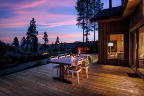 Une terrasse en bois avec une table et des chaises, d’où l’on peut admirer un paysage montagneux au crépuscule.
