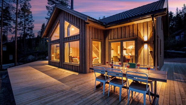 Une cabane en bois dotée de grandes fenêtres, d’une terrasse et d’un coin repas avec des chaises et une table.