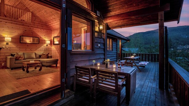 Un chalet confortable avec un intérieur en bois, un coin repas avec une table et des chaises, et un balcon donnant sur un paysage boisé.