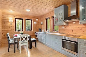 Interior de una cabaña de madera con una cocina que incluye estufa, armarios y una mesa de comedor con sillas.