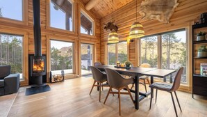 A spacious wooden interior with a large dining table, chairs, a fireplace, and large windows offering a view of the forest.