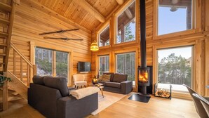 A log cabin interior with a fireplace, wooden floors, and large windows offering views of the forest.