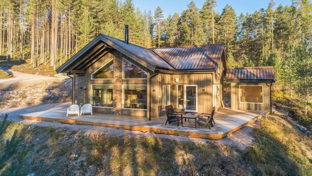 En modern timmerstuga med en stor veranda, stora fönster och en skog som bakgrund.