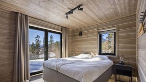 A bedroom with a large bed, a nightstand, a window with a view of trees and mountains, and wooden walls. - Lothlórien Lodge 8p - Four Bedroom Chalet, Sleeps 8 (Vrådal)