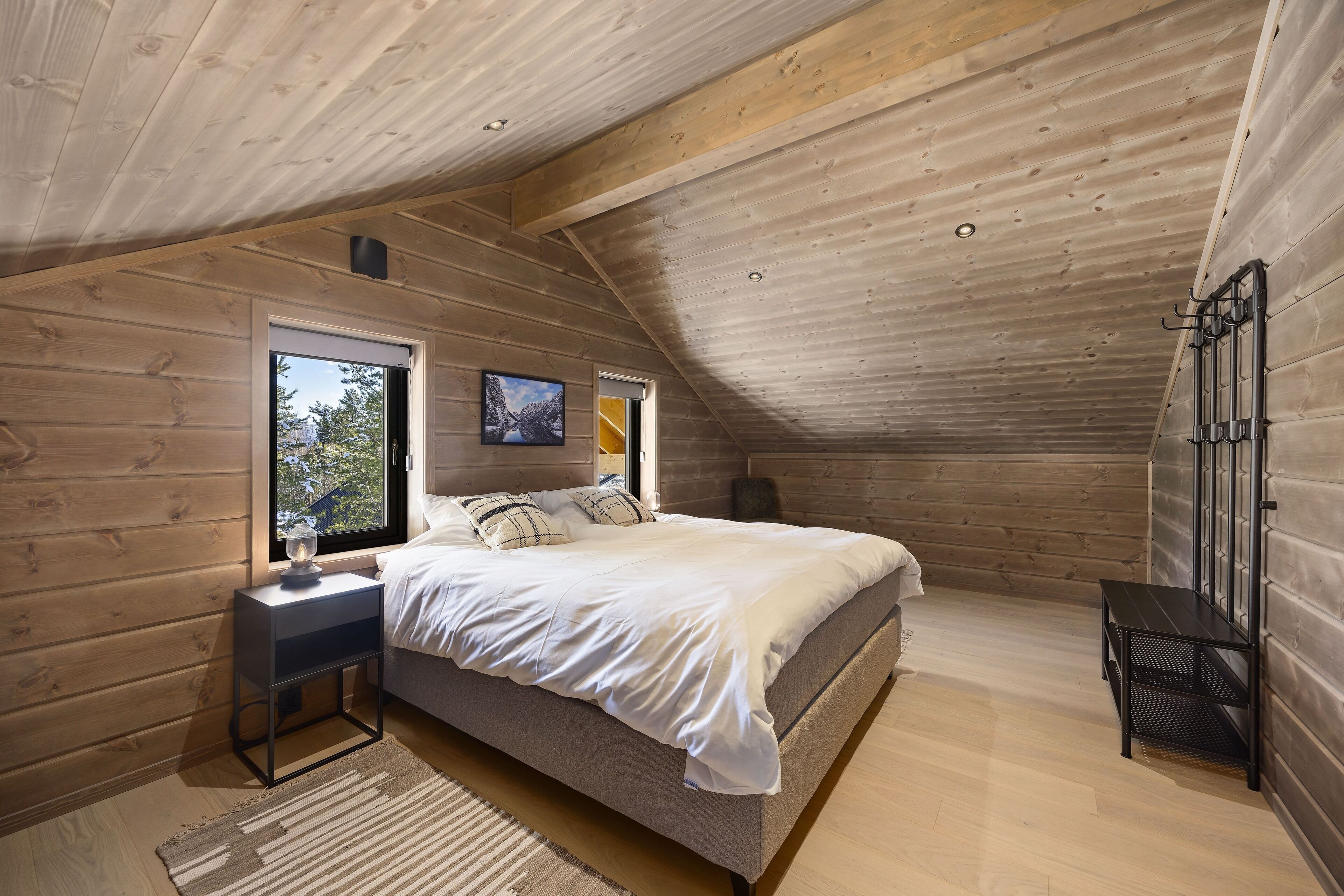 Un dormitorio con techo inclinado, paredes de madera, una cama, una mesita de noche, un banco y una ventana con vistas a árboles.