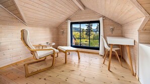 Een kamer met houten lambrisering en een schuin dak, met een groot raam met uitzicht op bergen, en een kleine witte tafel met een stoel.