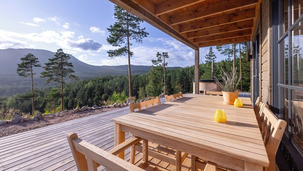 Terraza de madera con una mesa y bancos, con vistas a una cadena montañosa boscosa.