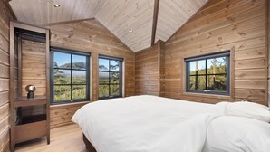 Un dormitorio con cama, dos ventanas, techo de madera y vistas a un bosque y montañas.