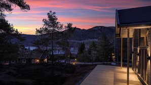 Une terrasse en bois mène à un bâtiment entièrement vitré, d’où l’on peut admirer un paysage montagneux au crépuscule.