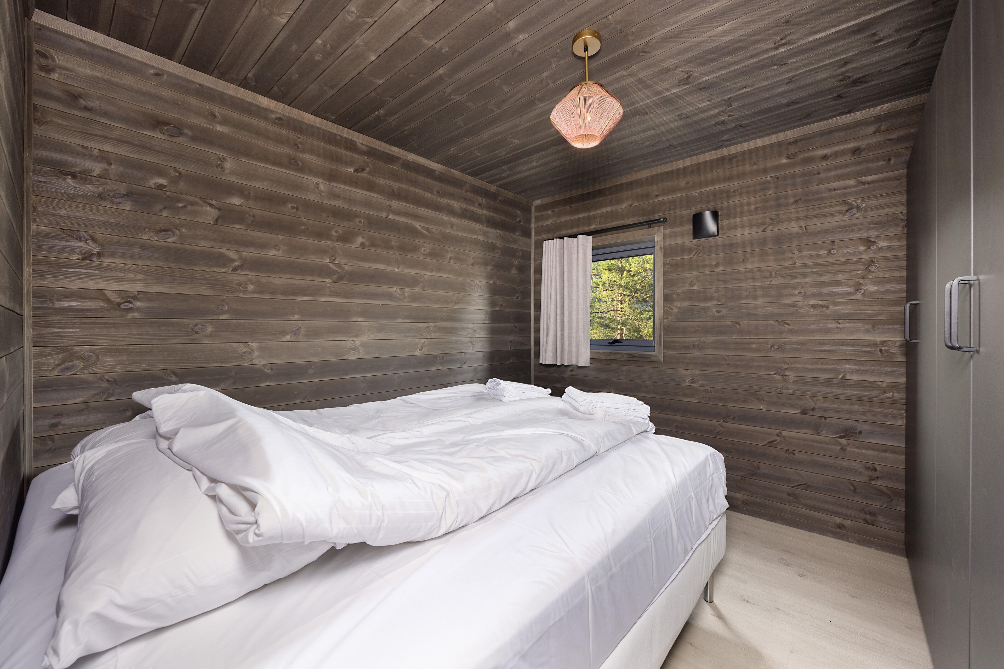 A bedroom with a wooden ceiling, a bed with white bedding, a window with a curtain, and a pendant light.