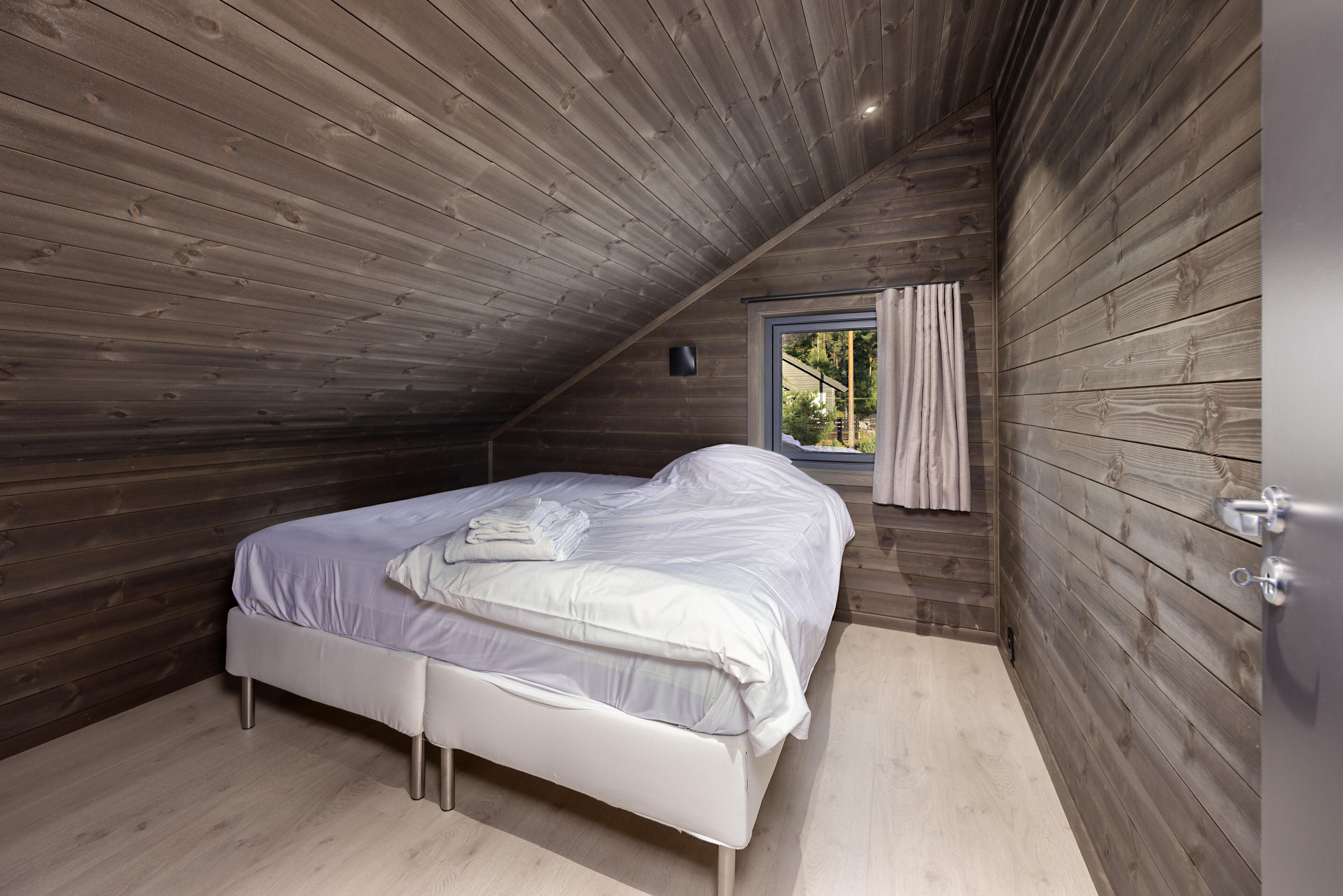 A bedroom with a wooden sloped ceiling, a bed with white bedding, a window with curtains, and a small wall-mounted light.