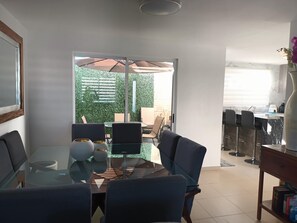 Ett modernt matområde med ett glasbord och stolar, utsikt över en grön utomhusyta och ett köksområde med barstolar.