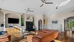 A spacious living room with a leather sofa, a flat-screen TV mounted on the wall, a fireplace, and a ceiling fan. - House in Bedford Woods (Placerville)