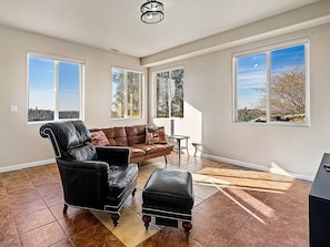 A spacious living room with a leather sofa, a matching ottoman, and a small side table. Large windows provide ample natural light and views of the outdoors.
