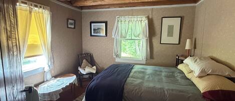 Ein Schlafzimmer mit Holzbalkendecke, einem Bett mit Kissen und Decke, einem Sessel mit Sitzkissen, einem kleinen Tisch mit Spitzenbettwäsche und einem Fenster mit Vorhängen.
