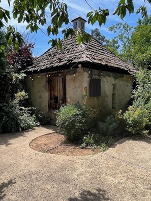 A rustic, single-story building with a thatched roof, surrounded by dense foliage and a gravel path.