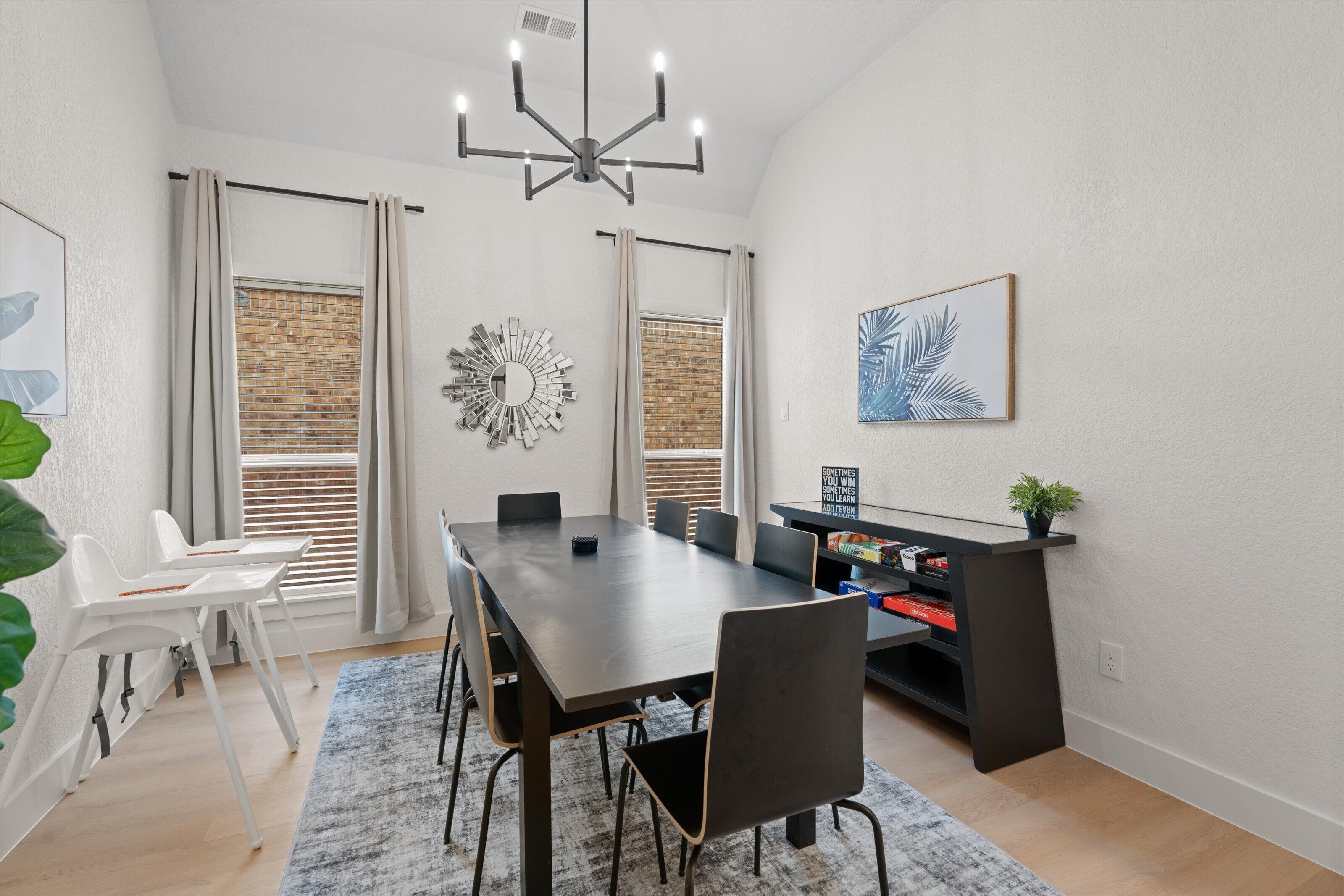Un espace de salle à manger moderne avec une table en bois foncé, des chaises noires et un lustre. On y trouve deux fenêtres équipées de stores, un miroir et une bibliothèque remplie de livres et ornée d’une plante.