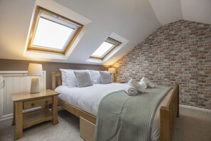 Ein Schlafzimmer mit schrägverdeckter Decke, zwei Dachfenstern, einer Ziegelwand, einem hölzernen Bettgestell, einem Nachttisch und einer Lampe.