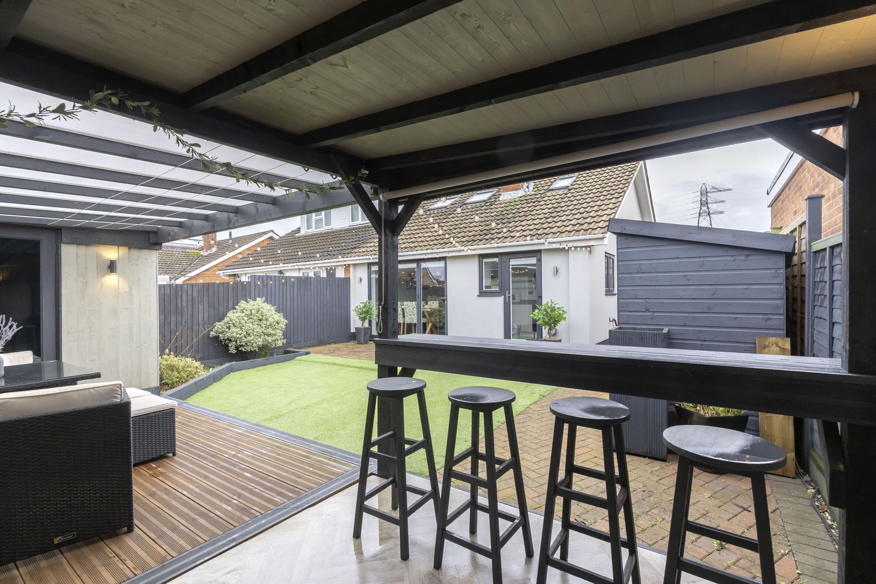 Une terrasse extérieure couverte avec une plateforme en bois, des tabourets de bar noirs et un coin salon comprenant une table et des coussins.