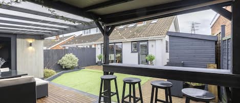A covered outdoor patio with a wooden deck, black bar stools, and a seating area with a table and cushions.