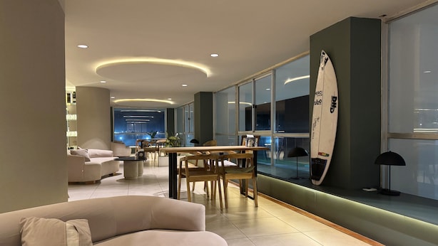 Ein modernes Wohnzimmer mit einer Couch, einem Esstisch und Stühlen. Ein Surfbrett lehnt an der Wand und eine Lampe steht auf dem Tisch.