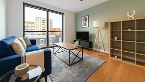 A modern living room with a blue sofa, a coffee table, a TV, a bookshelf, and a large window offering a view of buildings. - Luxury Studios | E8 4DN | Balcony (London)