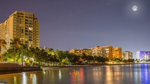Ein nächtliches Stadtbild mit einem hohen Hotelgebäude, Palmen und einer ruhigen Uferpromenade.
