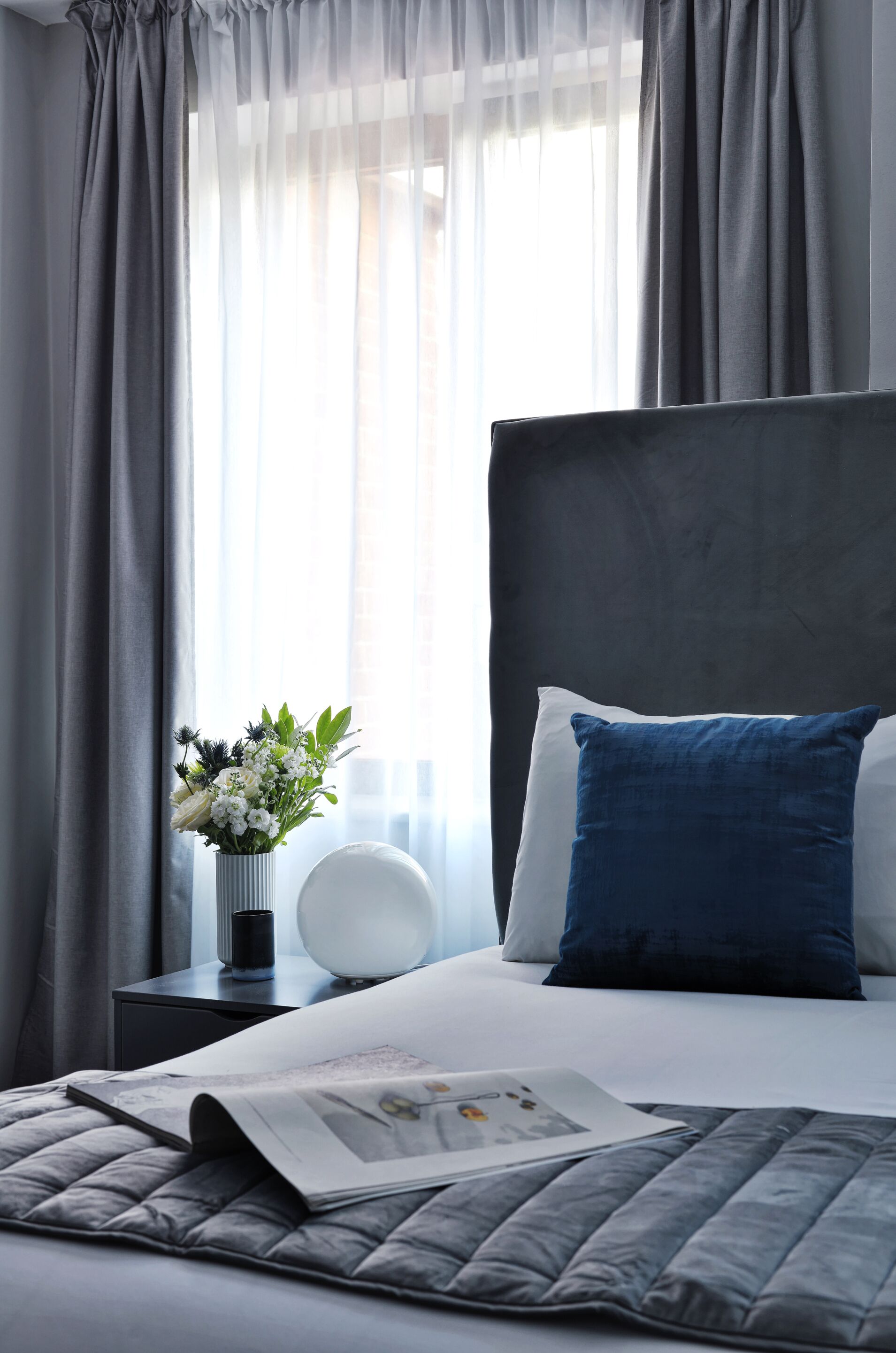 A neatly made bed with a blue pillow, a white pillow, and a gray quilt. There is a white vase with flowers on a bedside table.